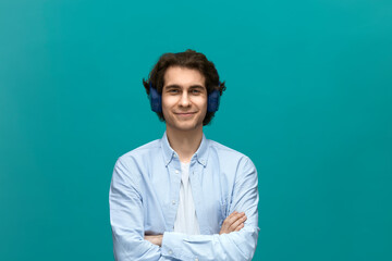 I am glad. Portrait of a young beautiful man wearing white t-shirt and blue shirt with headphones fold ones arms and looking at camera