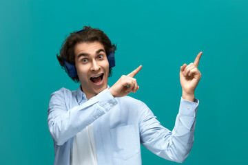 This is real shock. Portrait of a young beautiful man wearing white t-shirt and blue shirt in headphones. Point to something top at right by his forefingers