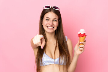 Young caucasian woman in swimsuit holding a cornet ice cream isolated on pink background points...
