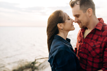Fototapeta premium Young man and woman in love hug at sunset near the sea, they gently touch each other's noses and smiling. Romantic mood