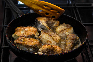 Pieces of fish are fried in a pan. A wooden spatula flips the fish. Cooking concept