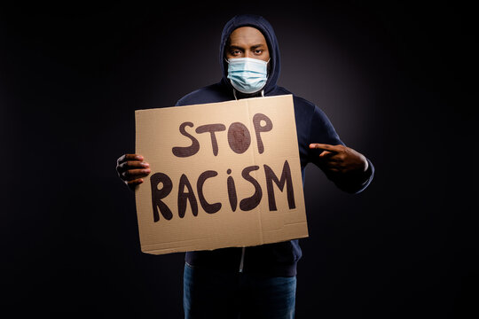 Photo of serious afro american guy hold point index finger card board banner share stop racism discrimination message wear jumper medical mask isolated black color background