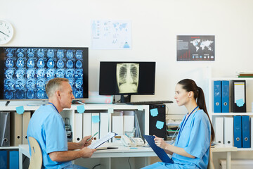 Obraz premium Side view portrait of two adult medics talking while sitting at workplace in modern clinic interior with CT scans in background, copy space