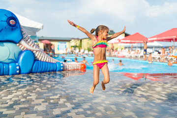 Little slim cute girl in bright swimsuit poses on backdrop of children's water zone in an open-air on warm summer day during vacation. Concept of children's entertainment and outdoor activities.