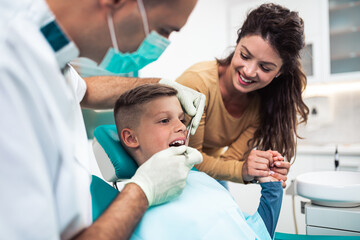 Cute little boy sitting on dental chair and having dental treatment. His mother is with him.