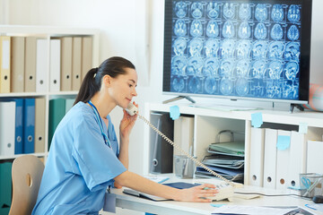 Side view portrait of young female medic speaking by phone while sitting at desk in hospital...