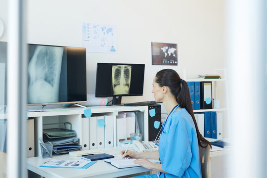 Side view portrait of young female medic looking at chest x-ray images and writing on clipboard while sitting at desk in clinic, copy space