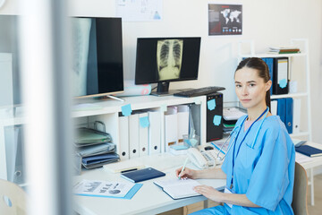 Waist up portrait of young female doctor looking at camera while posing at workplace with chest x-ray scans in background, copy space
