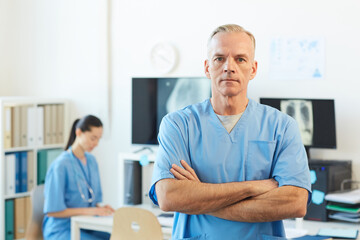 Waist up portrait of confident mature doctor looking at camera while standing with arms crossed in modern hospital interior, copy space