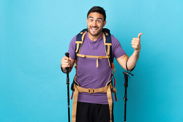 Young caucasian man with backpack and trekking poles isolated on blue background giving a thumbs up...