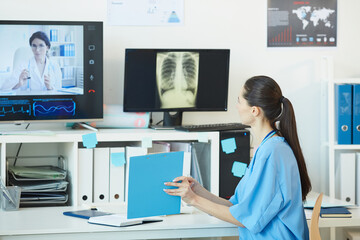Obraz na płótnie Canvas Back view at young female medic talking to doctor by video call and showing documents while sitting at workplace in office with chest x-ray images in background, copy space