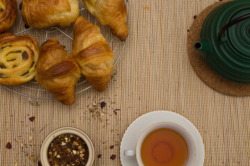 Breakfast or snack, tea with croissant, fine white cup with golden edges with small green kettle for tea, and small brown bowl with tea herbs, on neutral colored textured background.