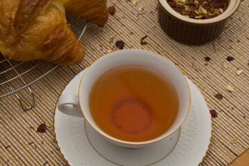 Breakfast or snack, tea with croissant, fine white cup with golden edges and small brown bowl with tea herbs, on neutral colored textured background.