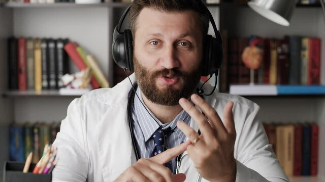 Doctor is talking to camera. Attractive bearded general practitioner doctor in white coat explains something while looking at camera