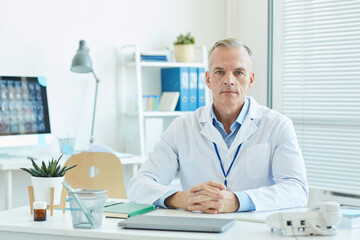 Portrait of mature male doctor wearing lab coat and looking at camera while sitting at desk in office of modern clinic, copy space
