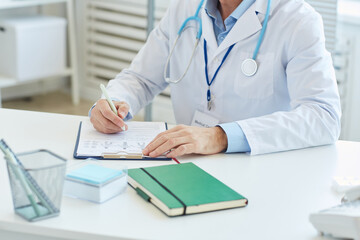 Fototapeta premium Portrait of unrecognizable doctor writing on clipboard and wearing lab coat while sitting at desk in office of modern clinic, copy space