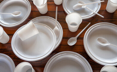 Platos, vasos y cubiertos de pástico blanco desechables esparcidos en una mesa de madera oscura.