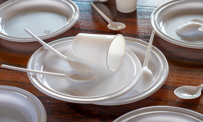 Platos, vasos y cubiertos de pástico blanco desechables esparcidos en una mesa de madera oscura.