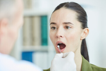 Unrecognizable male doctor taking swab sample from mouth and throat of young woman for virus test...