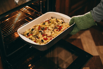 Home cooking in quarantine. Woman removes cooked, baked potatoes with mushrooms and spices from the oven.