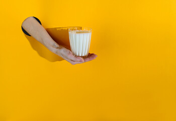 The hand from the hole holds a glass with kefir on a yellow background. Horizontal orientation,...