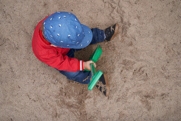 one-year-old boy with a cap, red jacket, blue pants and sneaker sits in the sand and digs with a green shovel