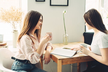 Two young girls spend time together at home, looking at a magazine and drinking coffee