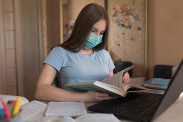 Obraz premium Teenager girl, home lessons at a laptop, medical mask, protection against bacteria viruses, reads a paper book textbook, video tutoring lessons, home education, preparation for college and school.