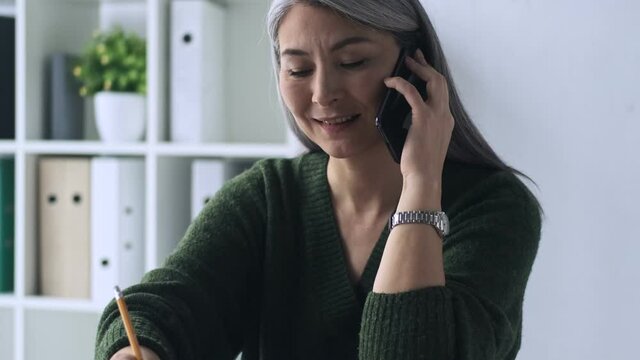 A positive pleased mature woman is making notes while talking on the smartphone working in the office