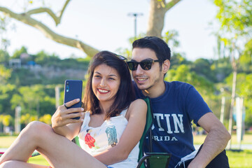 Portrait of cheerful couple man and woman 20s taking selfie photo while sitting on bench in green park