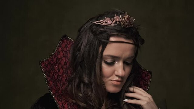 Halloween. Beautiful queen in cloak with crown at dark background. Portrait of young brunette vampiress. Close-up in 4K, UHD