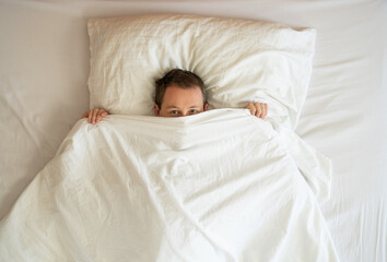 Image from above of young man hiding behind bed linens during the morning.