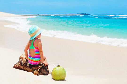 Little baby girl in hat sit on white sand beach. Child drink young coconut, look at ocean and enjoying. Kids travel lifestyle. Family summer holiday. Activity on tropical Hawaii and Caribbean islands.