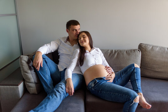 A man and a pregnant woman are sitting hugging on the couch. - Powered by Adobe