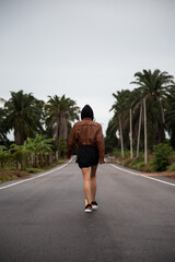 Fototapeta na wymiar Girl in a brown leather jacket is on a paved road with a yellow stripe amongst the palm trees