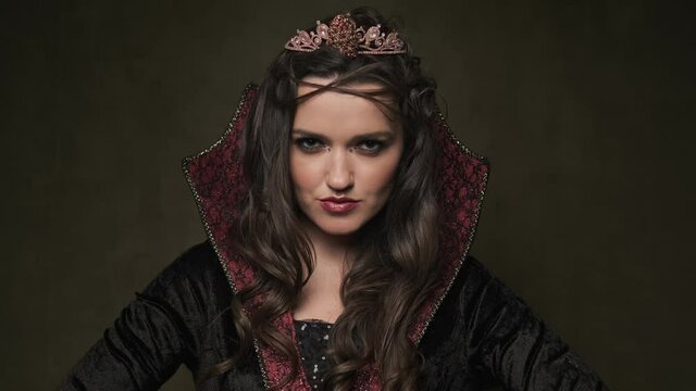 Female in cloak and crown. Beautiful queen at dark background. Portrait of vampiress looking at camera. Close-up shot in 4K, UHD