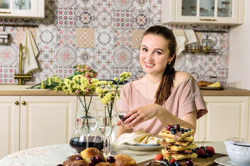 A young woman sits at a dining table in the kitchen with a cup of tea in her hands.