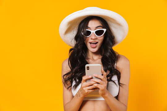 Image of stylish surprised woman in swimsuit and hat using cellphone