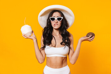 Image of displeased young woman in swimsuit and hat holding coconuts