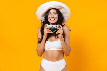 Image of happy beautiful woman in swimsuit and hat using retro camera