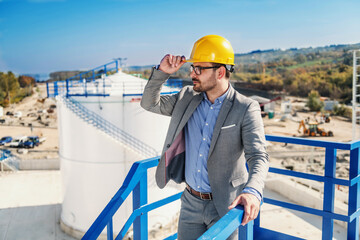 Young proud refinery owner in suit with helmet on head looking around and holding helmet. In...