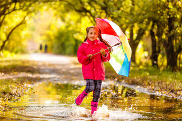 Very cute little girl in pink Jacket and rubber boots is jumping over a puddle on a rainy day