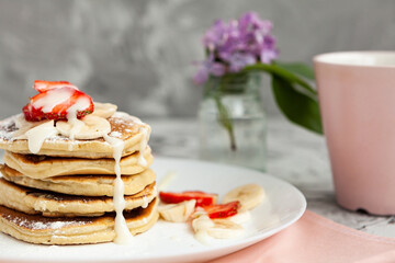 A stack of pancakes with a pink tea mug and  fresh strawberries on a white plate on a light background. A small bouquet of lilacs in a glass vase stands next to the Breakfast.