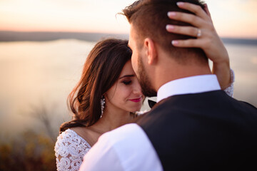The bride and groom are hugging on the background of the lake during sunset.