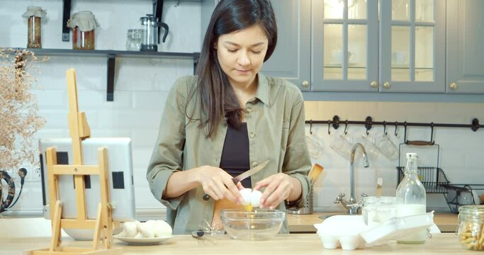 Young attractive woman preparing dough for pancakes or cookies using a tablet for tips in the kitchen. Reading the recipe online, technology devices, cooking process.