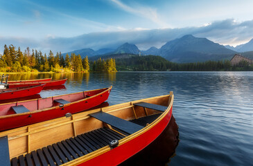 Fototapeta premium Beautiful sunny landscape. View on mountain lake with crystal clear azure water in High Tatras. Slovakia. Red Boats on the water glowing in sunlight at sunset. Awesome Autumn landscape. Strbsce pleso