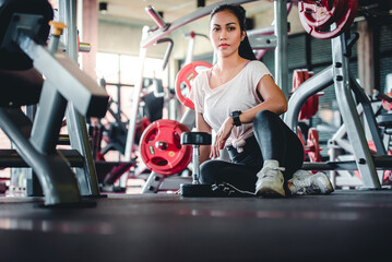 Asian women with beautiful tan skin sit and relax at the exercise bench. After sports training with dumbbells For health in the fitness center Concept: sporty girls sitting in the gym