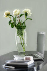 glass vase with white peonies stands on the working place with an open laptop
