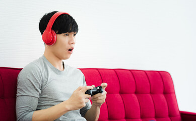 Young Asian man sitting on red couch acting excited and playing console game in living room