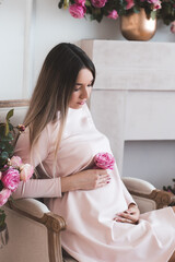 Young pregnant woman 20-25 year old holding fresh pink rose flower sitting in chair in room....